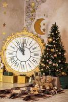 Stylish xmas living room interior with Christmas tree decorated with candles, garland. Big gold white clock, many gift boxes and present toys indoors. Decoration stars and moon on wall. New Year photo