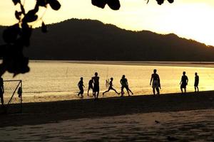 silueta, grupo, de, gente, jugar al fútbol, en la playa, en, el, ocaso, con, seascape, y, montaña, en, plano de fondo foto