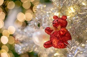 Selective focus on the right eye of the teddy bear doll decorated on the Christmas tree with glittering bokeh lights in background