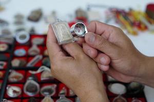 Closeup hands hold Buddha amulet and magnifying glass to check details and figure of object. Concept, faith and belief for Buddhist for holy and luck. Colleting amulet as hobbies, exchange, selling. photo