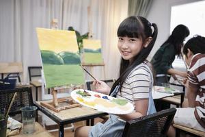 Portrait of Asian girl looking at camera and smiling with acrylic color picture painted on canvas in art classroom and creative learning with talents and skill at elementary school studio education. photo