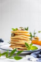 Stack of fluffy pancakes with orange jam, blueberries, coconut chips and mint, light background. Traditional breakfast. High key photography.