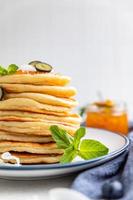 Stack of fluffy pancakes with orange jam, blueberries, coconut chips and mint, light background. Traditional breakfast. High key photography.