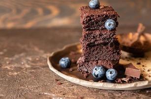 Chocolate brownie squares decorated with blueberry and pieces of dark chocolate and cocoa powder on craft ceramic plate, brown background.