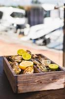 una variedad de ostras frescas con lima y limón en una caja de madera. mariscos frescos terraza de café al aire libre. fondo borroso con vista al club náutico. foto