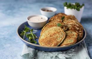 Vegetable fritters or pancakes made of zucchini, broccoli or spinach on plate with microgreen and dip, blue background. Healthy vegetarian food. photo