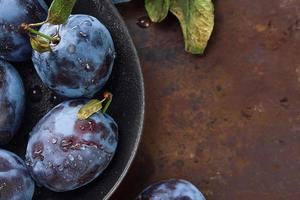 Close-up of ripe plums with leaves in a black plate on an old table, horizontal background, top view. Beautiful ripe prunes, harvesting fruits in autumn, eco products from the farm photo