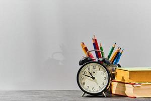 concepto de regreso a la escuela o idea del día del maestro. bolígrafos, lápices, libros, un despertador sobre la mesa, en el fondo de una pizarra gris con espacio para copiar. foto