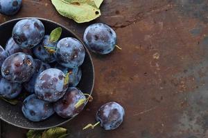 Fresh plums with leaves in a black plate on an old table, horizontal background, top view with copy space. Beautiful ripe prunes, harvesting fruits in autumn, eco products from the farm photo