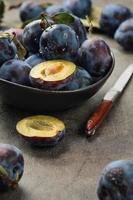 Selective focus, shallow depth of field. Ripe plums with leaves and halves of plums in a ceramic bowl on a gray table, next to a knife, close-up selective focus. Harvesting prunes in autumn. photo