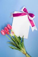 ramo de tulipanes rosas y papel para texto con lazo lila sobre fondo azul. feriado, san valentín, día internacional de la mujer y la madre, 8 de marzo, cumpleaños. copie el espacio foto