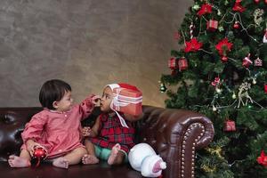 dos diversos amigos bebés sonríen felizmente mientras juegan con el presente en el sofá vistiéndose con traje de navidad y sombrero de santa con árbol de navidad en la parte posterior para el concepto de celebración de temporada foto