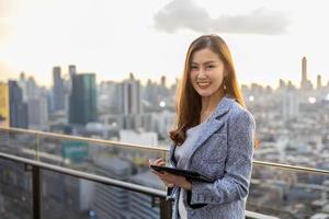 joven mujer de negocios asiática en traje formal con tableta digital de pie fuera del edificio de rascacielos para el concepto de bienes raíces, vivienda y desarrollo urbano foto