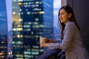joven asiática disfrutando de la vista nocturna desde su balcón mientras mira el paisaje urbano de rascacielos por la noche con una taza de café caliente para la vivienda y el concepto de estilo de vida moderno foto