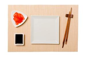 Empty white square plate with chopsticks for sushi and soy sauce, ginger on brown sushi mat background. Top view with copy space for you design photo