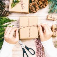 Woman wrapping christmas present , girl prepares xmas gifts with fir tree and pine cone. Hand crafted gift on wooden background with Christmas decor. Top view, copy space. Toned photo