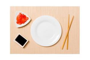 Empty oval white plate with chopsticks for sushi, ginger and soy sauce on brown sushi mat background. Top view with copy space for you design photo