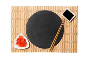 Emptyround black slate plate with chopsticks for sushi and soy sauce, ginger on yellow bamboo mat background. Top view with copy space for you design photo