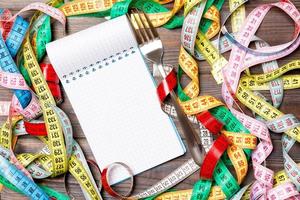 Group of colorful measure tapes, open notebook and fork on wooden background with empty space for your idea. Top view of healthy lifestyle concept photo