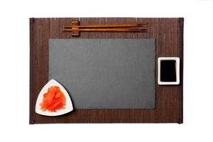 Empty rectangular black slate plate with chopsticks for sushi, ginger and soy sauce on dark bamboo mat background. Top view with copy space for you design photo