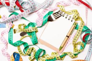 Top view of notebook with fork surrounded with colorful measuring tapes on white wooden background. Copy space with diet planning photo