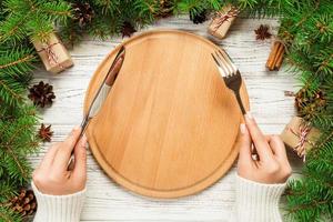 top view girl holds fork and knife in hand and is ready to eat. Empty wood round plate on wooden christmas background. holiday dinner dish concept with new year decor photo