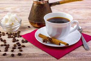 White cup of black hot coffee on saucer, served with coffee beans, white sugar cubes in a bowl, tea spoon, anise and cinnamon sticks on wooden textured background