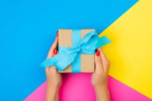 woman hands holding gift wrapped and decorated with blue bow on blue, pink and yellow background with copy space photo