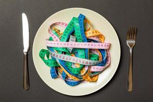 Top view of colorful measuring tapes on plate in the form of spaghetti with knife and fork on black background. Weight loss and diet concept photo