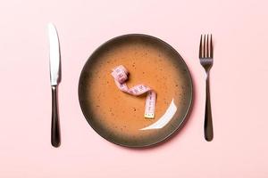 Round plate with measure tape inside with fork and knife on pink background. Top view of obesity concept photo