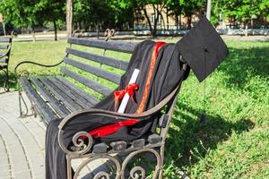 gorra de graduación, sombrero con borla negra, manto con un grado de papel en un banco del parque foto