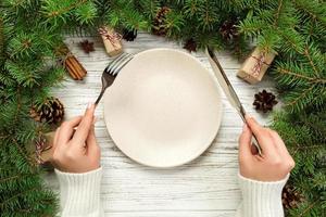 top view girl holds fork and knife in hand and is ready to eat. Empty plate round ceramic on wooden christmas background. holiday dinner dish concept with new year decor photo