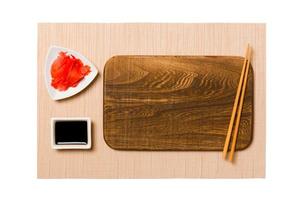 Empty rectangular brown wooden plate with chopsticks for sushi, ginger and soy sauce on brown sushi mat background. Top view with copy space for you design photo