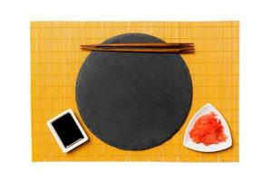 Emptyround black slate plate with chopsticks for sushi, ginger and soy sauce on yellow bamboo mat background. Top view with copy space for you design photo