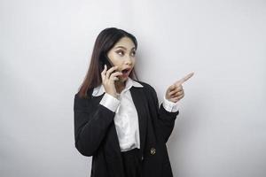 Shocked Asian businesswoman wearing a black suit pointing at the copy space beside her while talking on her phone, isolated by a white background photo