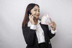 una joven empresaria feliz lleva traje negro, sostiene su teléfono y dinero en rupias indonesias aislada de fondo blanco foto