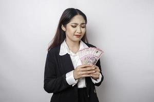 A confused young businesswoman is wearing black suit and holding cash money in Indonesian rupiah isolated by white background photo