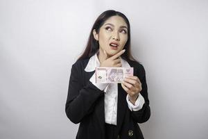 A thoughtful young woman is wearing black suit and holding cash money in Indonesian rupiah isolated by white background photo