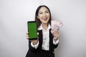 A happy young businesswoman is wearing black suit, showing her phone and money in Indonesian rupiah isolated by white background photo