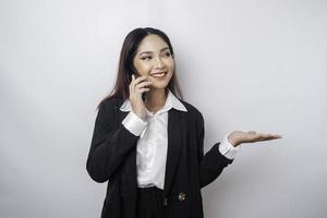 emocionada mujer de negocios asiática con traje negro apuntando al espacio de copia a su lado mientras habla por teléfono, aislada de fondo blanco foto