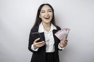 A happy young businesswoman is wearing black suit, holding her phone and money in Indonesian rupiah isolated by white background photo