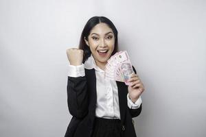 una joven mujer de negocios asiática con una expresión feliz y exitosa que usa traje negro y tiene dinero en rupias indonesias aislada de fondo blanco foto