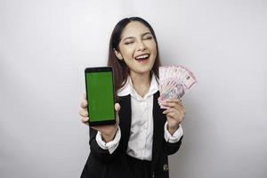 una joven y feliz mujer de negocios lleva traje negro, mostrando su teléfono y dinero en rupias indonesias aislada de fondo blanco foto