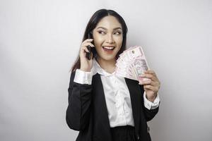 una joven empresaria feliz lleva traje negro, sostiene su teléfono y dinero en rupias indonesias aislada de fondo blanco foto