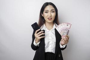 A confused young businesswoman is wearing a black suit, holding her phone and money in Indonesian rupiah isolated by a white background photo