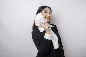 A thoughtful young woman is wearing black suit and holding cash money in Indonesian rupiah isolated by white background photo