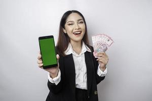 A happy young businesswoman is wearing black suit, showing her phone and money in Indonesian rupiah isolated by white background photo