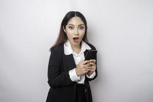 Surprised Asian businesswoman wearing black suit holding her smartphone, isolated by white background photo