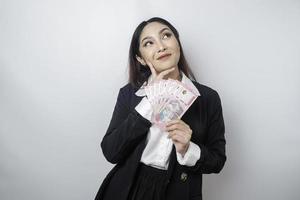 A thoughtful young woman is wearing black suit and holding cash money in Indonesian rupiah isolated by white background photo