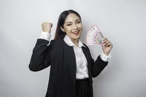 una joven mujer de negocios asiática con una expresión feliz y exitosa que usa traje negro y tiene dinero en rupias indonesias aislada de fondo blanco foto